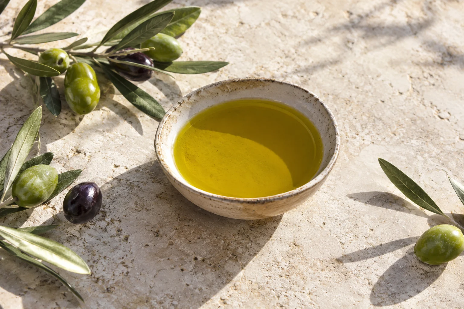 A ceramic bowl of olive oil with olives and leaves on limestone